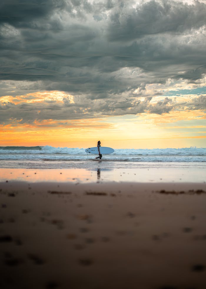 A serene view of a surfer walking along the beach with a surfboard at sunset in Essaouira, Morocco.